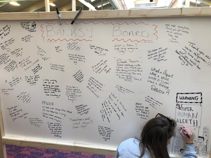 A whiteboard filled with short greetings from many of the staff. An artists' hand, botom right, can be seen drawing lots of people, cartoon-style.