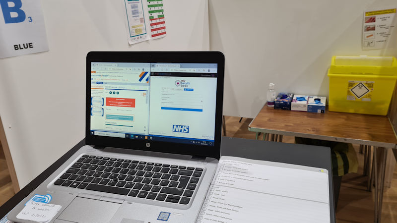 A laptop is in the foreground. Behind it you can see a sharps bin and other clinical paraphernalia in a small booth