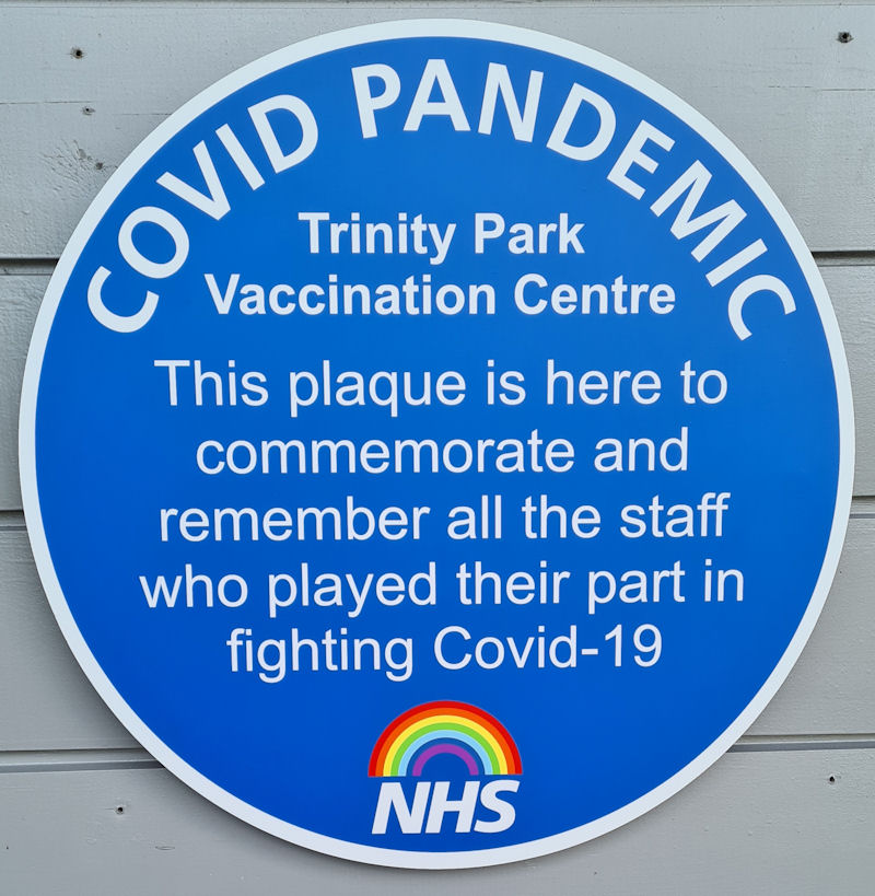 A round blue plauqe with the words: COVID Pandemic. Trinity Park Vaccination Centre. This plaque is here to commemorate and remember all the staff who played their part in fighting Covod-19. An NHS logo and rainbow is at hte bottom