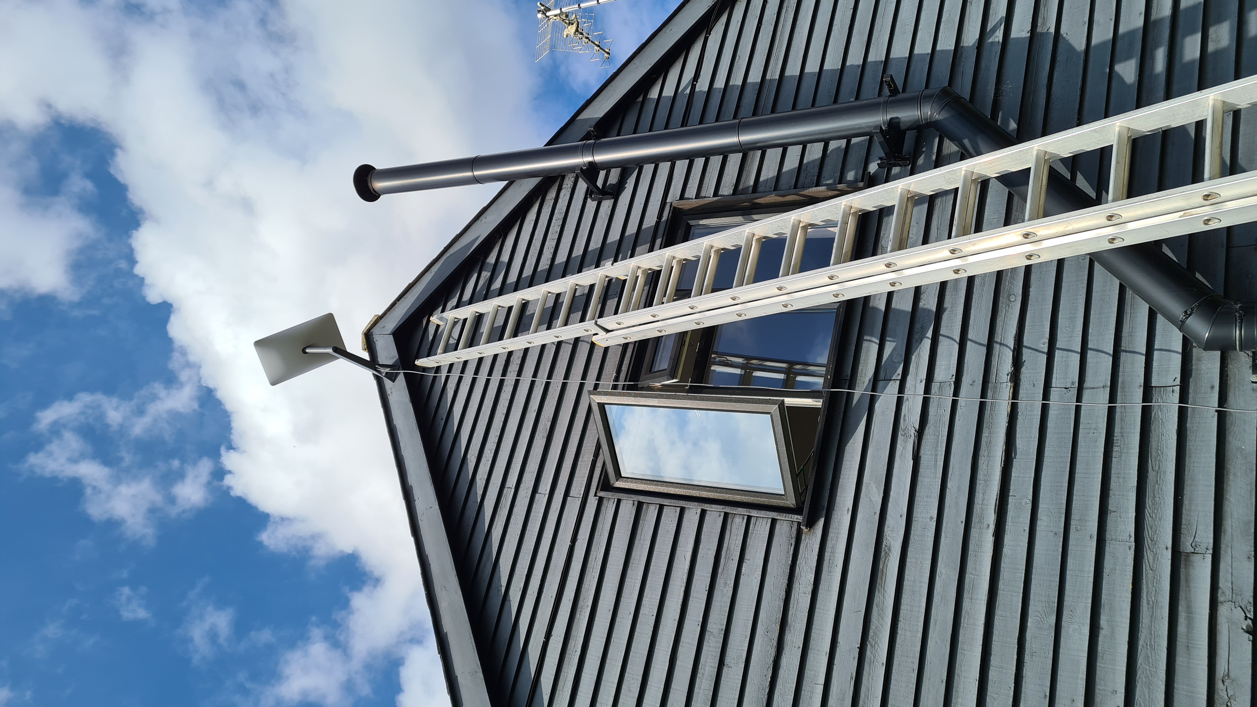 A tall ladder reaching up the gable end of a house, at the top of which is the Starlink stellite receiver