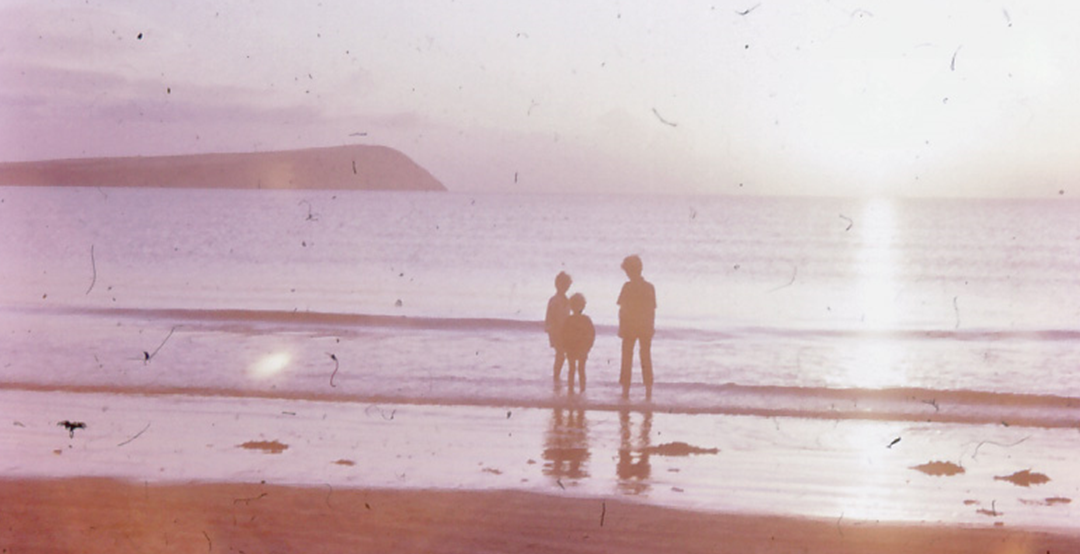 An obviously old photograph shows a beach at sunset with three boys silhouetted. In the background, a distinctive headland can be seen