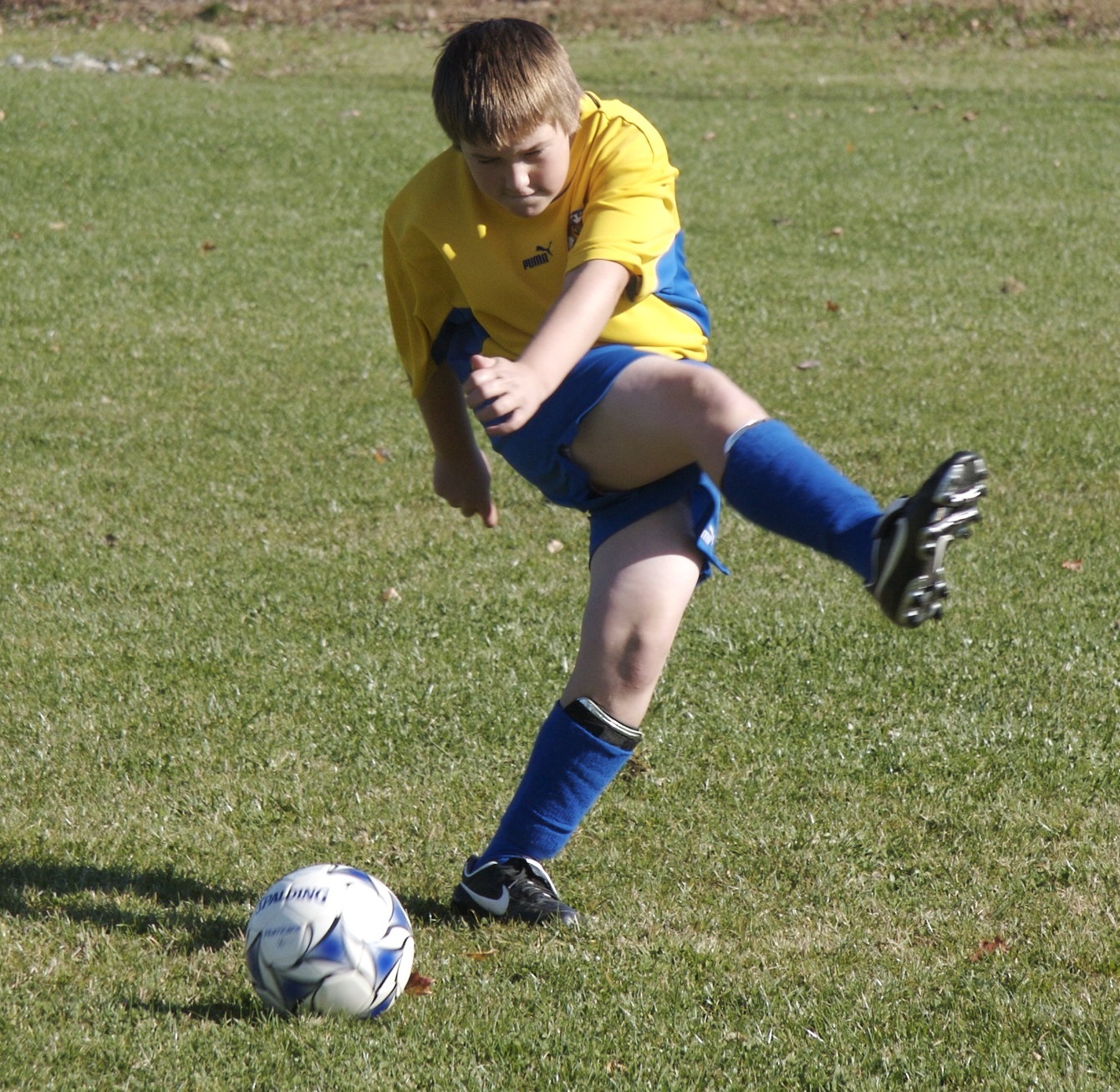 A boy of about 12 years old wearing a yellow and blue football kit kicks a ball