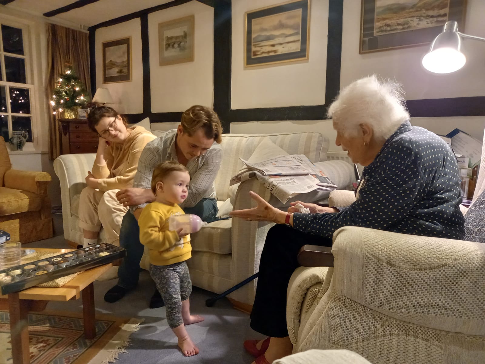 A 14 month-old , bottle in hand, approaches an elderly lady who has her arms out to greet her