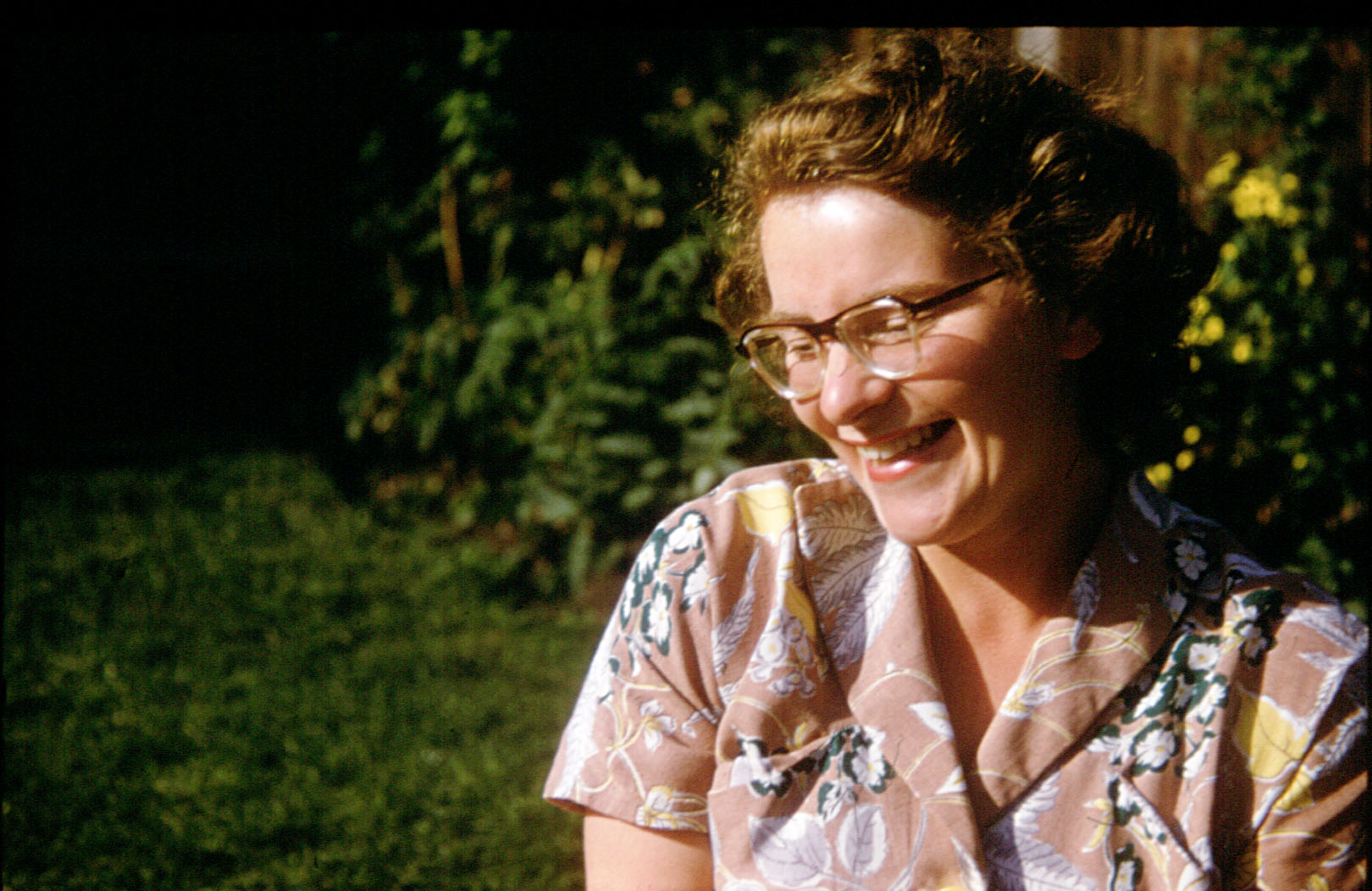 A woman in her 20s, with short hair looking down to her right and smiling. Her short-sleeved dress has a floarl pattern.